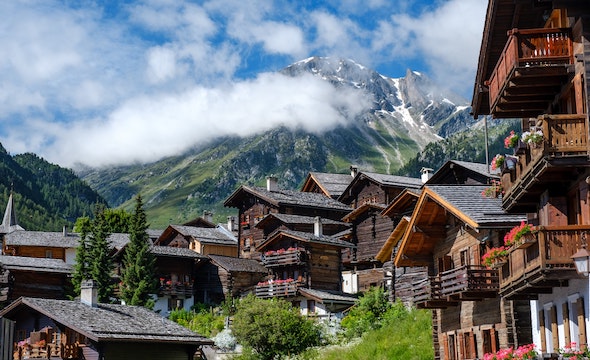 A picture of cozy wood Swiss chalets with balconies built into green hills covered in trees reaching up into snow peaked mountains that rise above thin wispy clouds.
