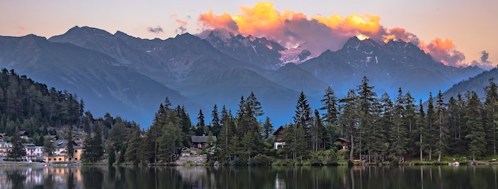 A picture of body of water reflecting cozy looking buildings and tall trees along the waters edge. With large Swiss mountains in the background topped with bright clouds reflecting the setting sun and warm colored evening sky.